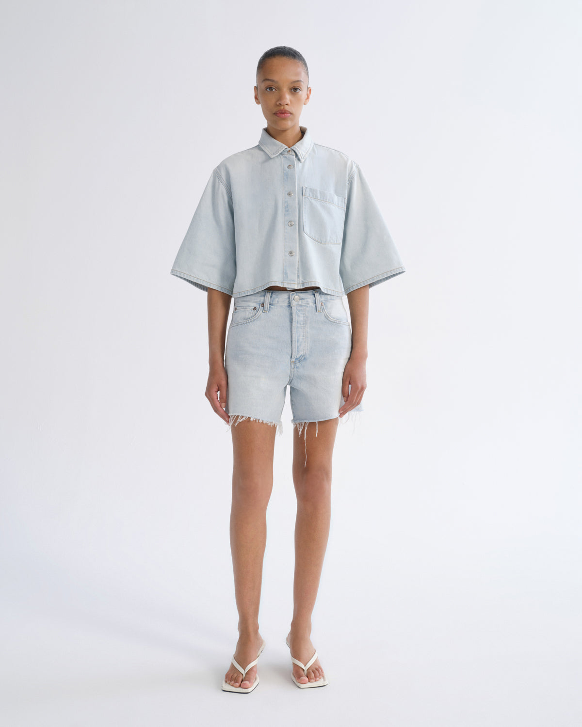 Woman wearing light blue denim shorts with a light blue short sleeve button-up shirt on a grey background.