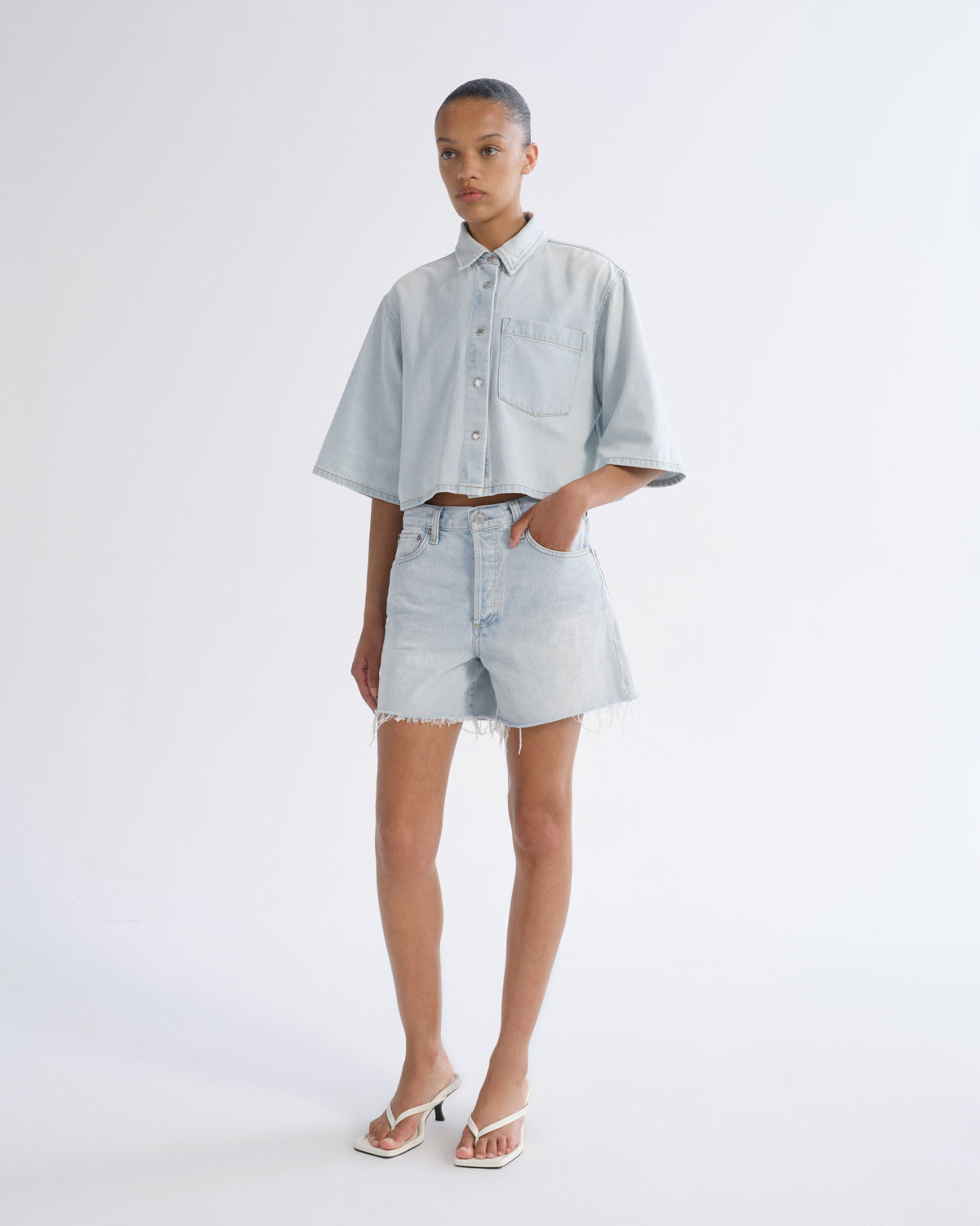 Woman wearing light blue denim shorts with a light blue short sleeve button-up shirt on a grey background.