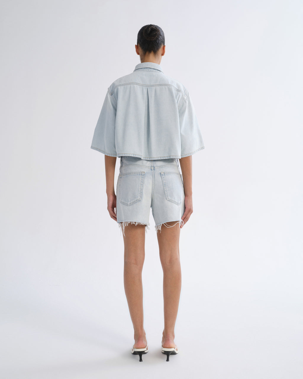 Woman wearing light blue denim shorts with a light blue short sleeve button-up shirt viewed from the back on a grey background.
