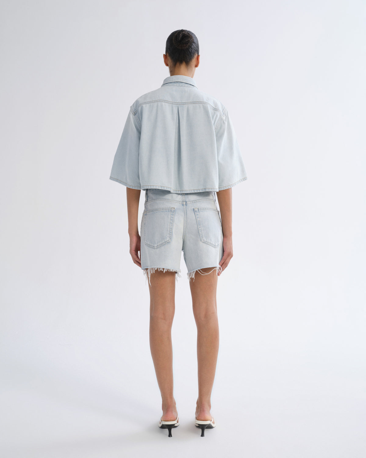Woman wearing light blue denim shorts with a light blue short sleeve button-up shirt viewed from the back on a grey background.