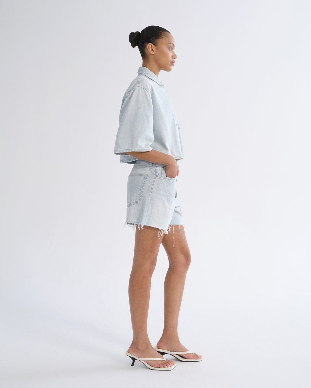Woman wearing light blue denim shorts with a light blue short sleeve button-up shirt viewed from the side on a grey background.