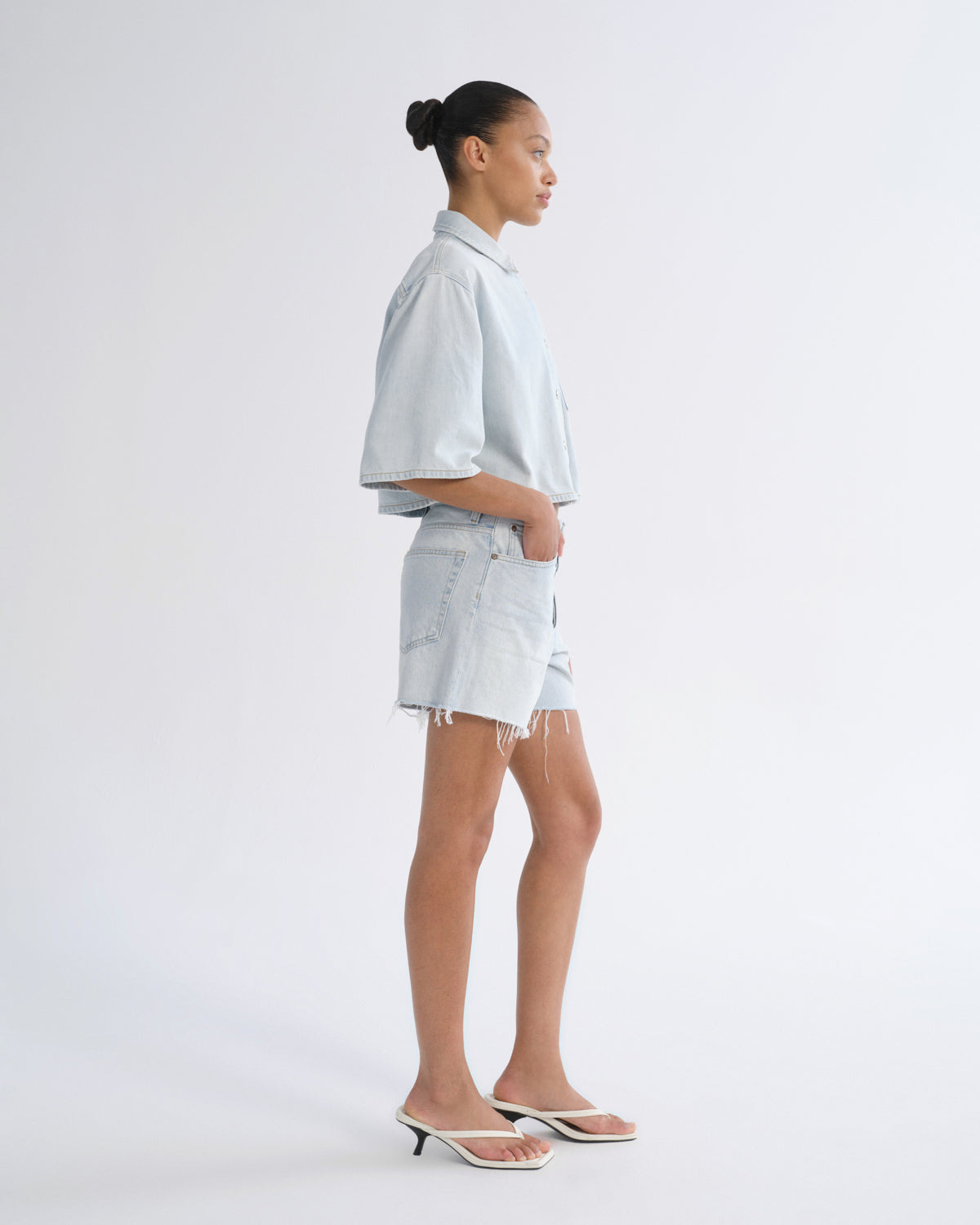 Woman wearing light blue denim shorts with a light blue short sleeve button-up shirt viewed from the side on a grey background.