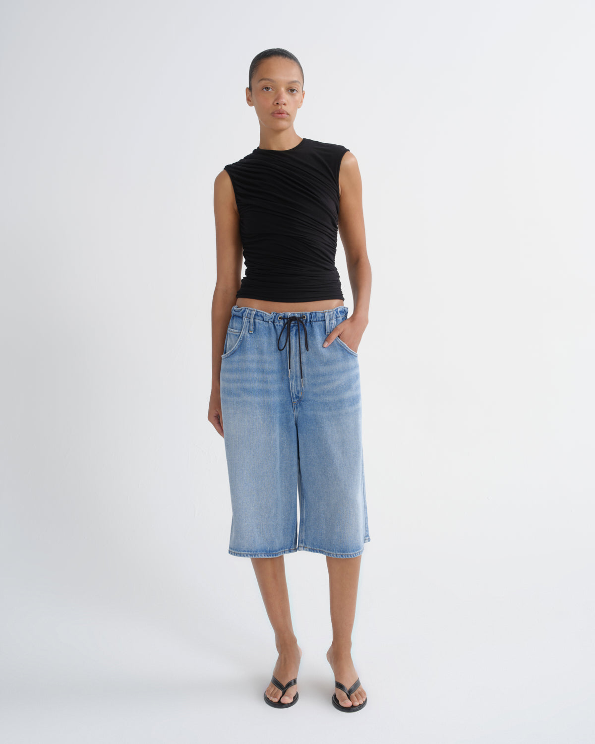 A model stands against a light gray studio background wearing a sleeveless black ruched top, light-wash blue denim cropped pants with a drawstring waist, and black flip-flop sandals.