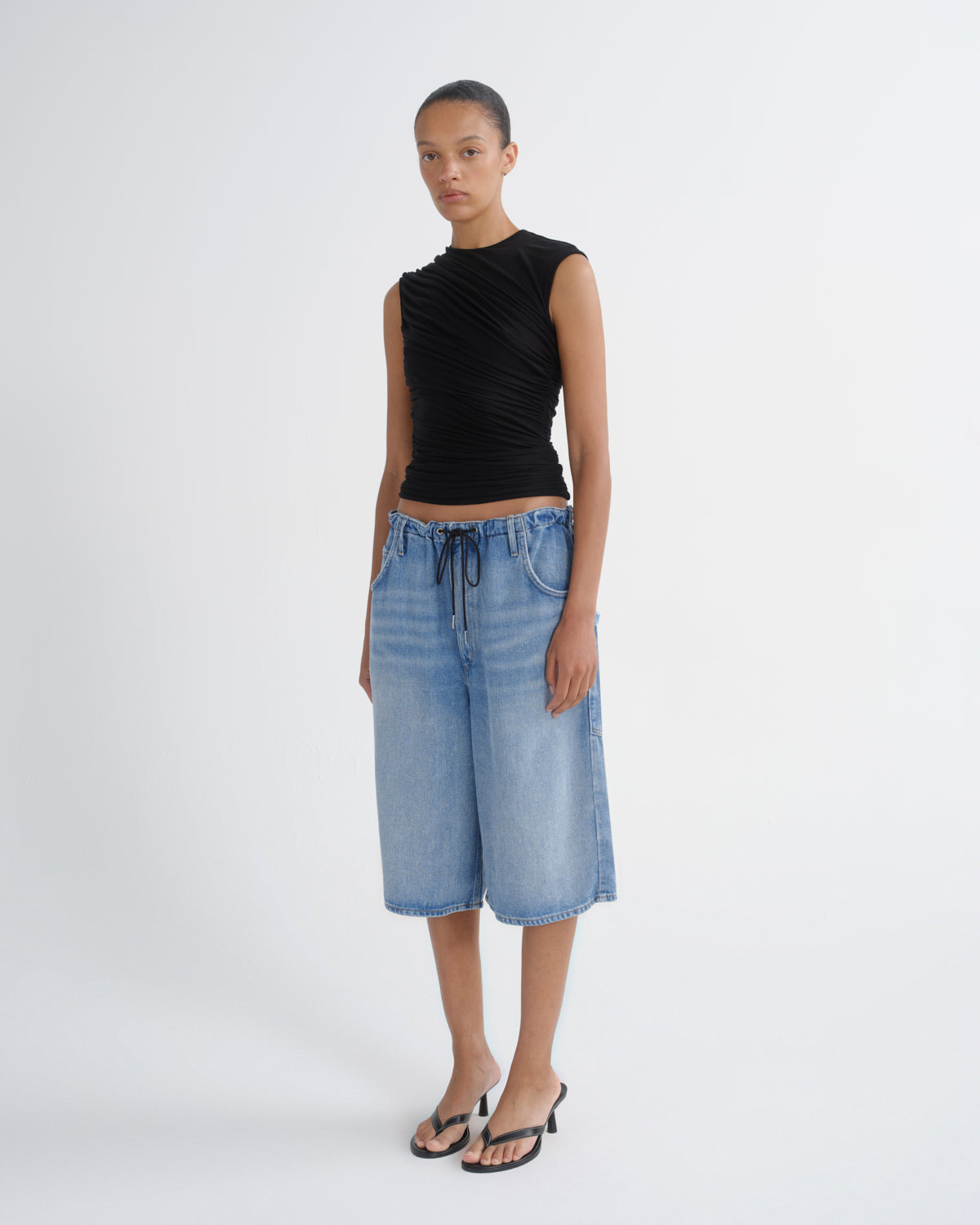 A model stands against a light gray studio background wearing a sleeveless black ruched top, light-wash blue denim cropped pants with a drawstring waist, and black heeled sandals.