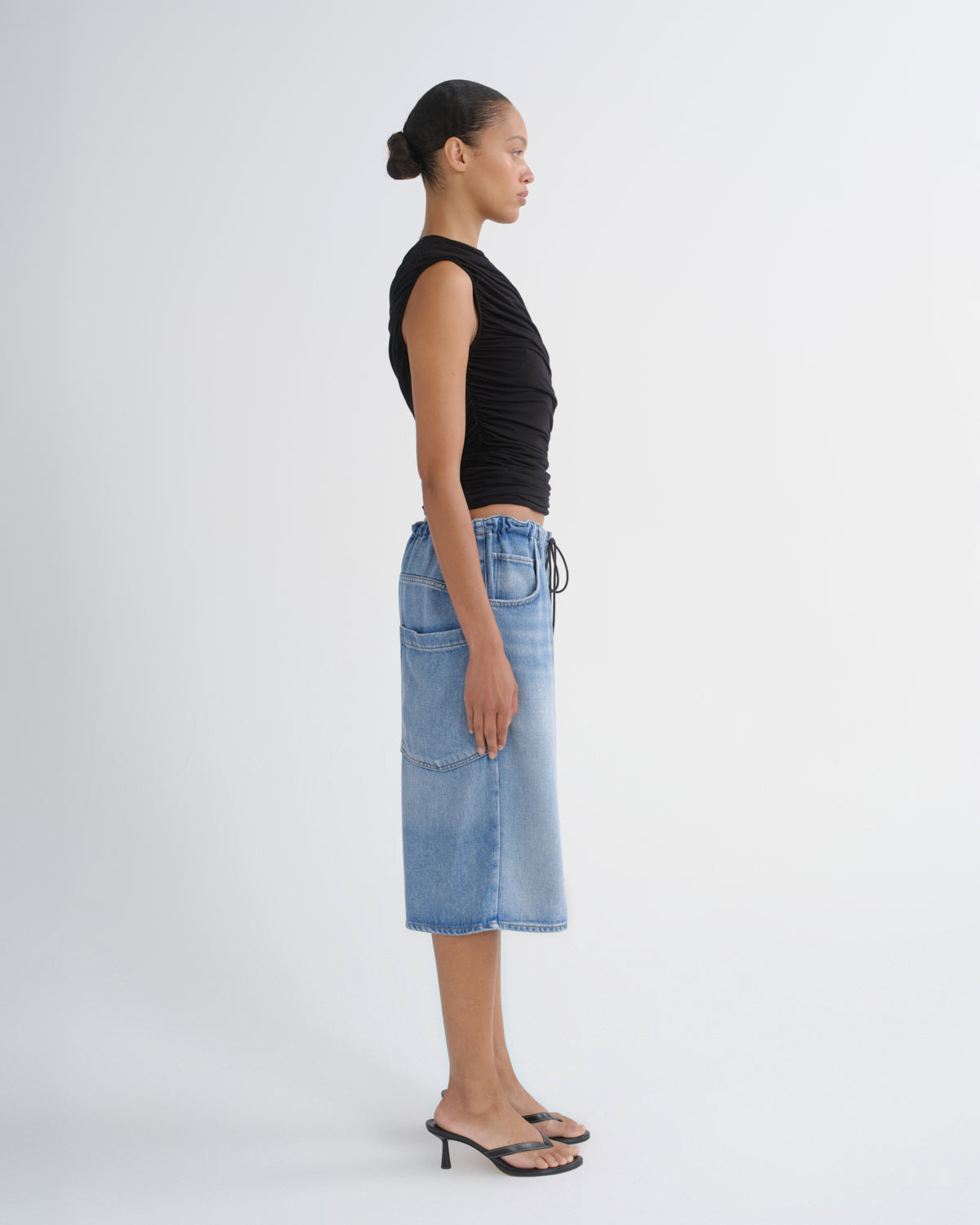 A model shown in profile wearing a black sleeveless top and a light-wash blue denim midi skirt with front patch pockets, standing against a plain light gray studio background.