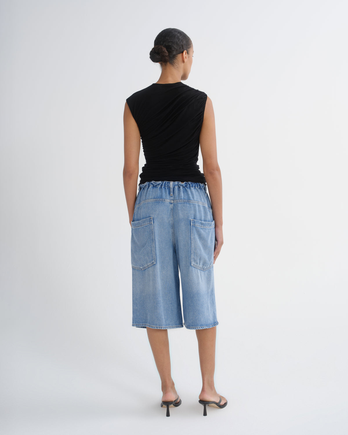 A model shown from the back wearing a black sleeveless top and light blue denim cropped pants with back patch pockets, paired with black heeled sandals, against a plain light gray studio background.