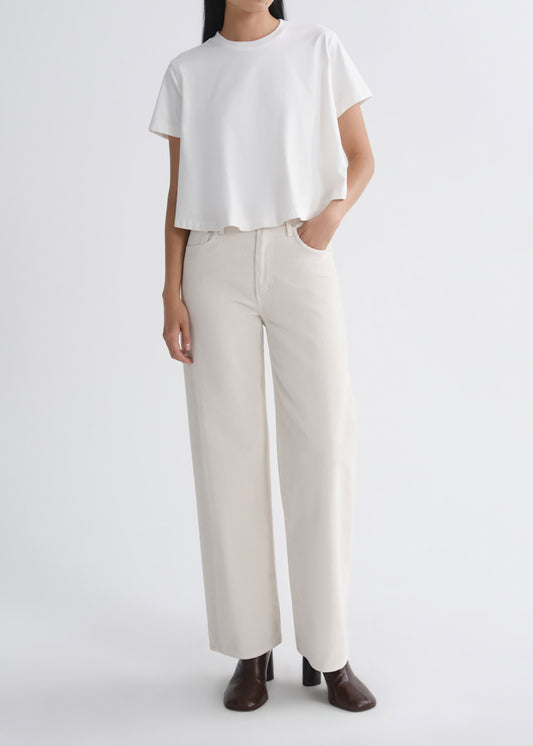 A model wearing a white short-sleeve cropped t-shirt and off-white wide-leg trousers, paired with dark brown heeled shoes, against a grey background.