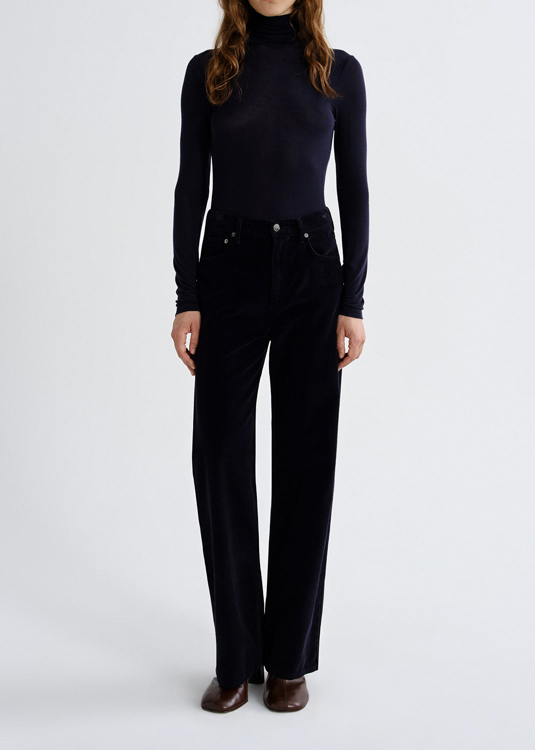 A model wearing a dark navy long-sleeve mock neck top and matching dark navy wide-leg trousers, paired with brown leather shoes, against a grey background.