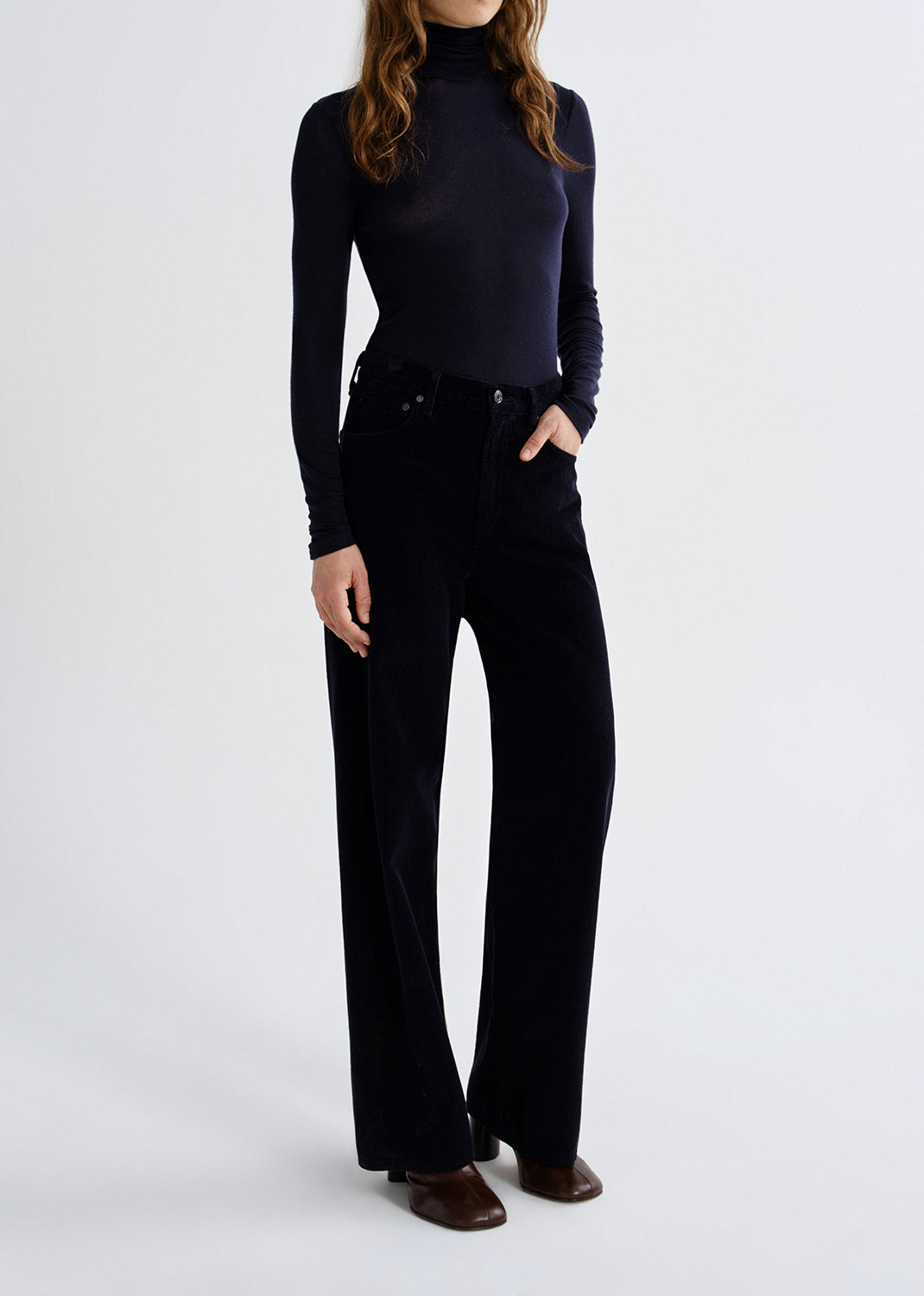 A model wearing a dark navy long-sleeve mock neck top and black wide-leg trousers, paired with brown leather shoes, against a grey background.