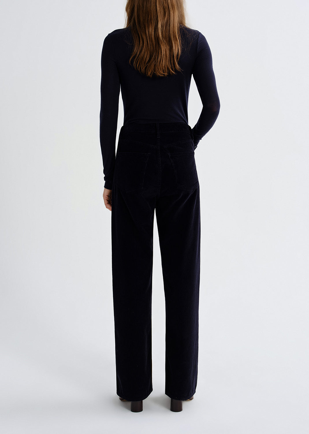 A woman viewed from the back wearing a dark navy long-sleeve top and matching wide-leg trousers against a grey background.