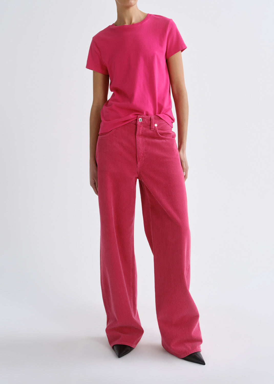 A model wearing a bright pink short-sleeve crewneck t-shirt and matching wide-leg jeans, styled with black pointed-toe heels against a grey background.