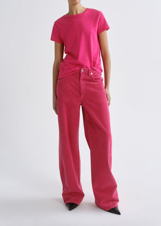 A model wearing a bright pink short-sleeve crewneck t-shirt and matching wide-leg jeans, styled with black pointed-toe heels against a grey background.