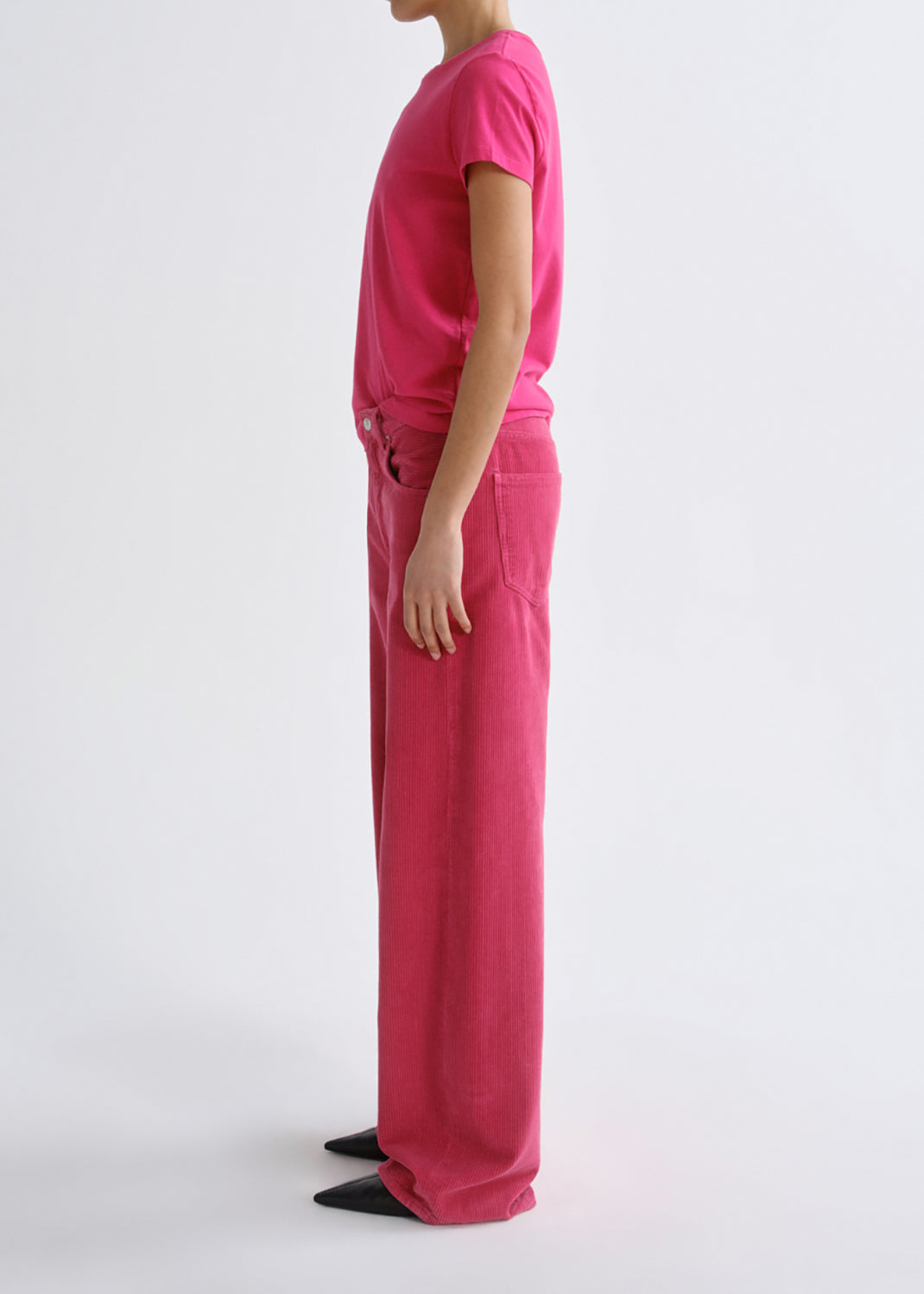 A model shown in profile wearing a bright pink short-sleeve t-shirt and matching wide-leg trousers, standing against a grey background.