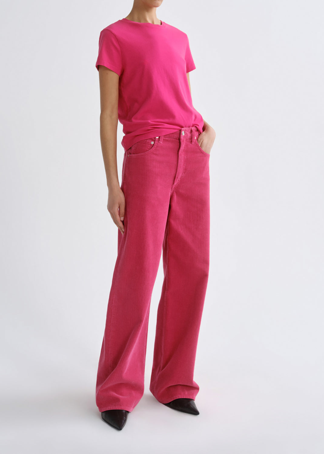 A model wearing a bright pink short-sleeve t-shirt and matching wide-leg corduroy trousers, paired with black pointed-toe heels, against a grey background.