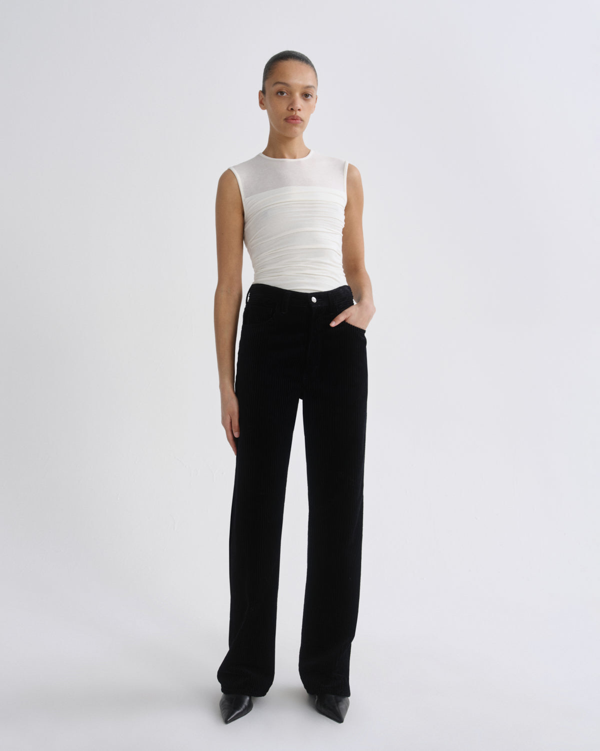 A model stands against a light gray studio background wearing a sleeveless white ribbed top and black wide-leg trousers.