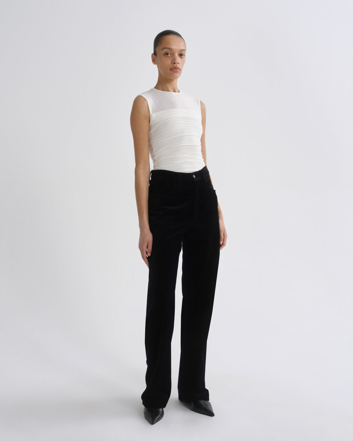 A model stands against a light gray studio background wearing a sleeveless white top with horizontal ruching and black trousers.