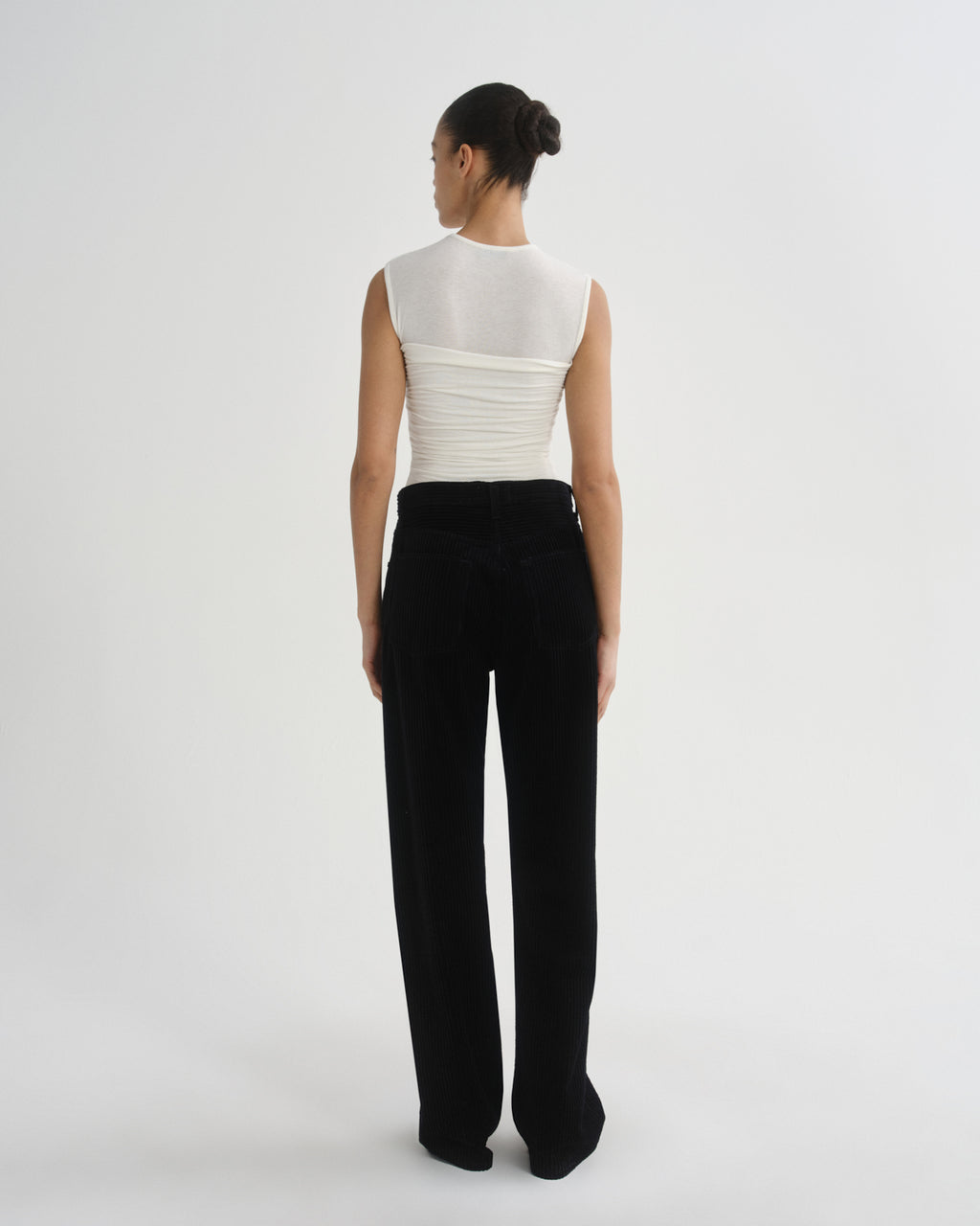 A model is shown from the back wearing a white sleeveless top with a sheer high neck and ruched back, paired with black wide-leg trousers, against a plain light gray studio background.