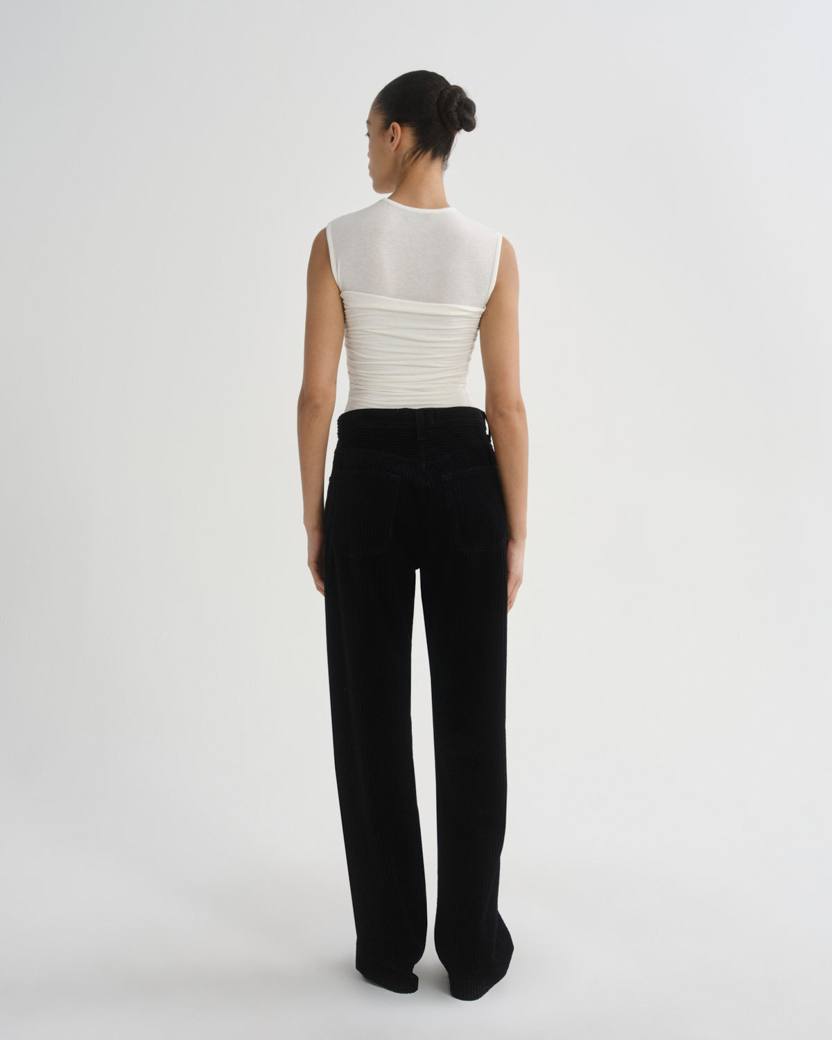A model is shown from the back wearing a white sleeveless top with a sheer high neck and ruched back, paired with black wide-leg trousers, against a plain light gray studio background.