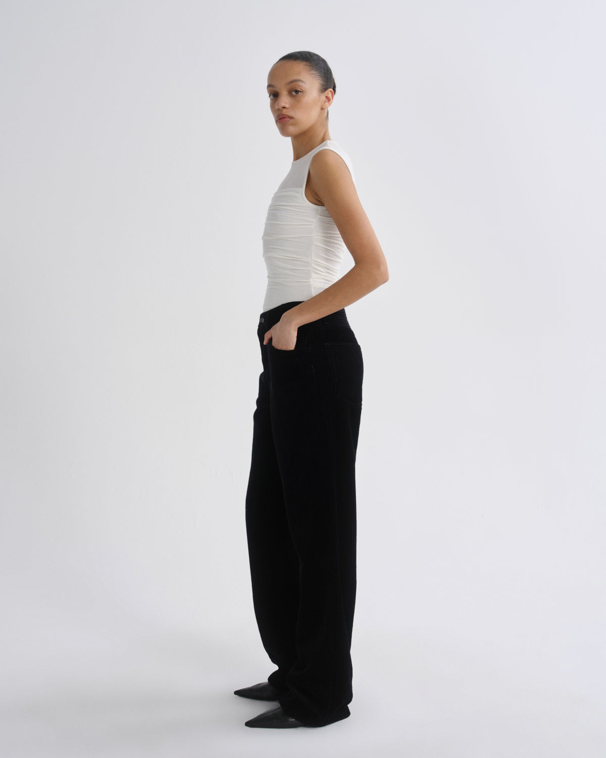 A model stands in profile wearing a white ribbed sleeveless top and black wide-leg trousers against a plain light gray studio background.