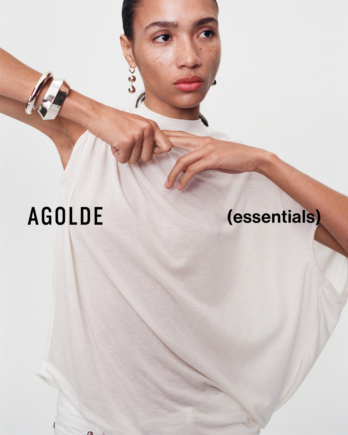 A model wearing a draped, oversized off-white sleeveless top with the text 'AGOLDE (essentials)' overlaid on the image, set against a plain light background.