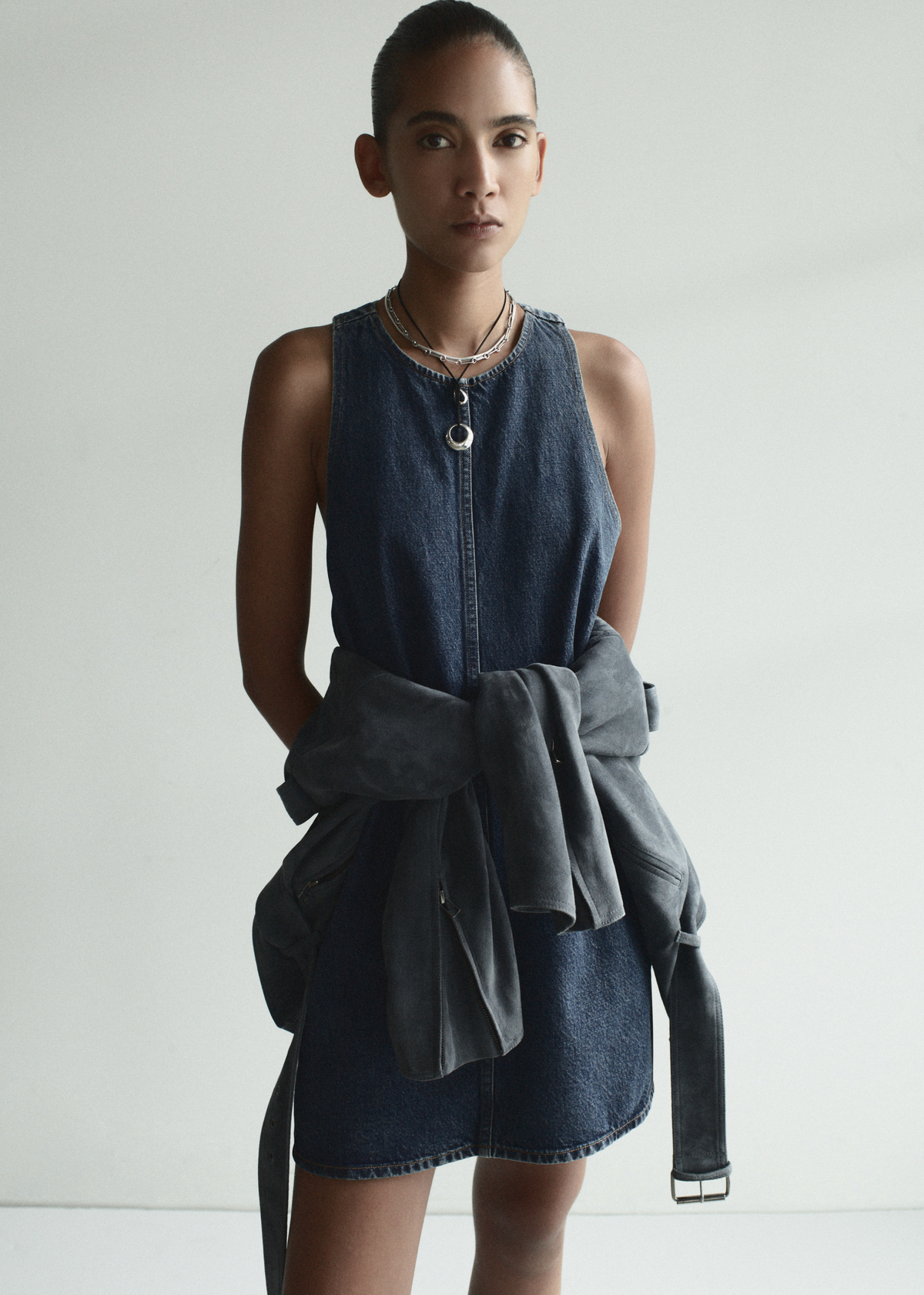 Woman wearing a denim dress with a gray jacket over her shoulder against a white background