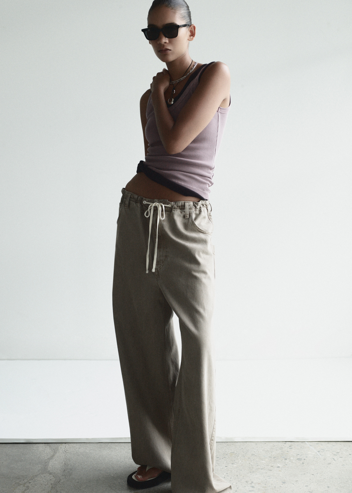 Person wearing a sleeveless top and beige pants with sunglasses on a plain background