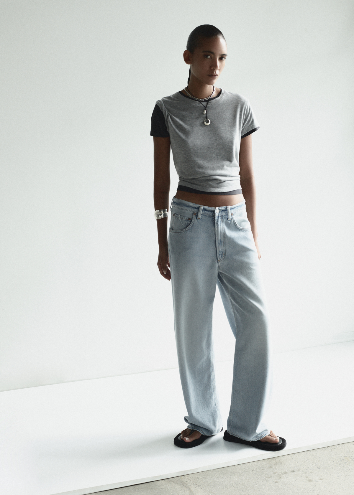 Person wearing a gray t-shirt and light blue jeans standing on a white floor with a white wall background