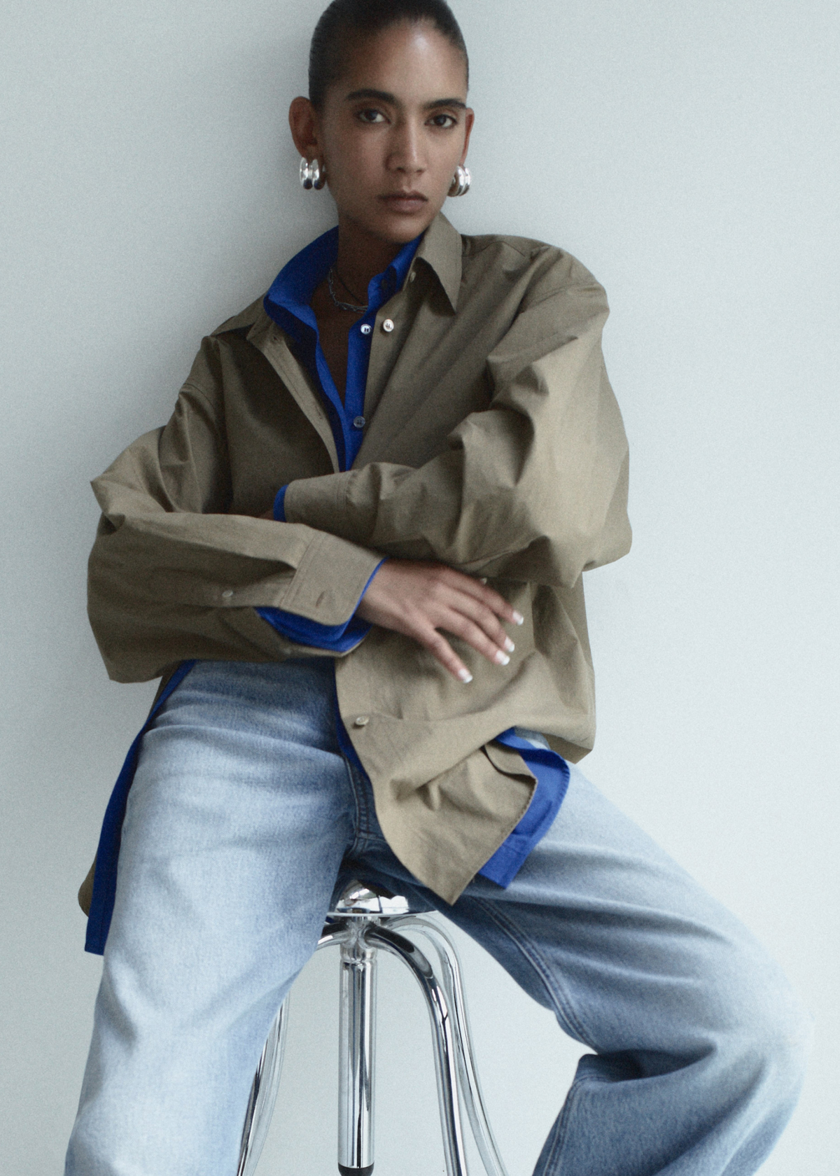 Person wearing a beige jacket with blue accents and light blue jeans, sitting on a clear stool against a white background.
