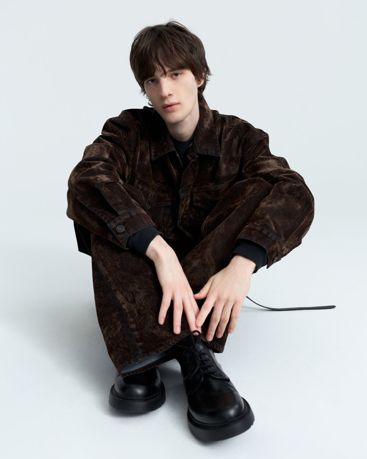A model wearing a dark brown textured corduroy jacket and matching trousers, paired with black leather boots, sitting against a light gray studio background.