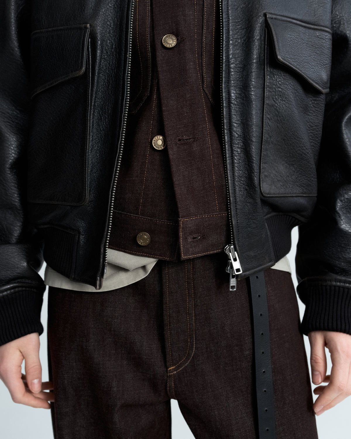 A close-up of a person wearing a black leather jacket over a dark brown corduroy vest and matching dark brown trousers, with a light beige shirt peeking out at the hem.