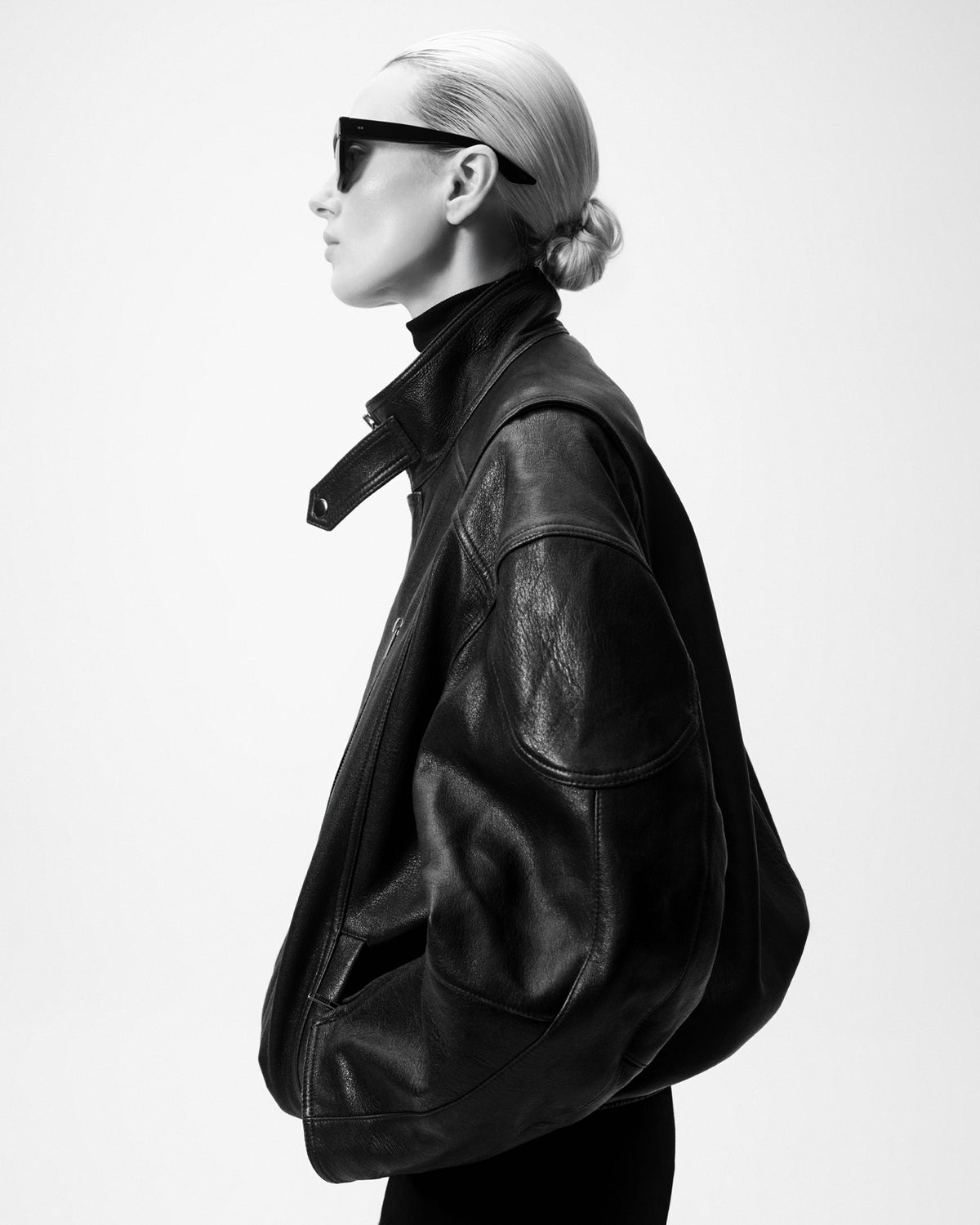 A black and white fashion photograph of a model in profile wearing a black leather jacket, black sunglasses, and a black turtleneck against a plain white background.