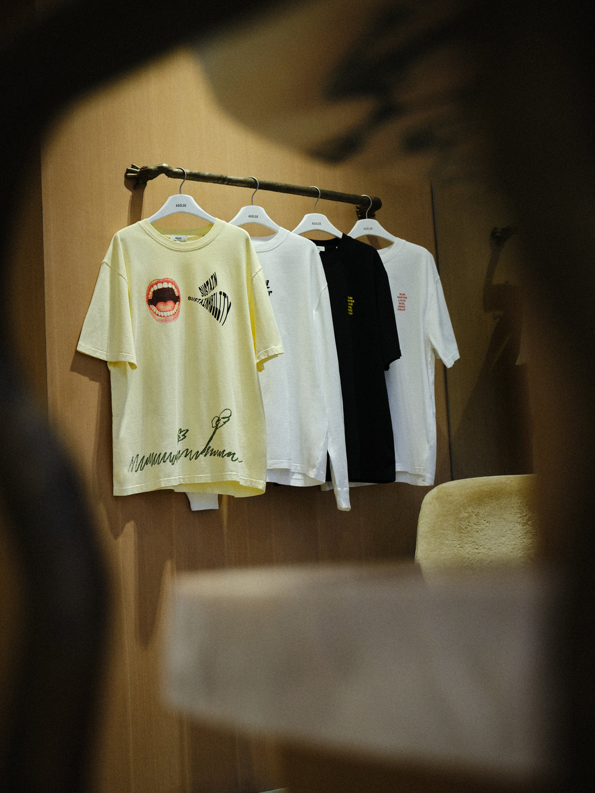 A row of hanging T-shirts on a clothing rack inside a store or room, with a pale yellow graphic tee in front, followed by white and black shirts. The scene is viewed through a dark foreground blur, and a chair is visible on the right.