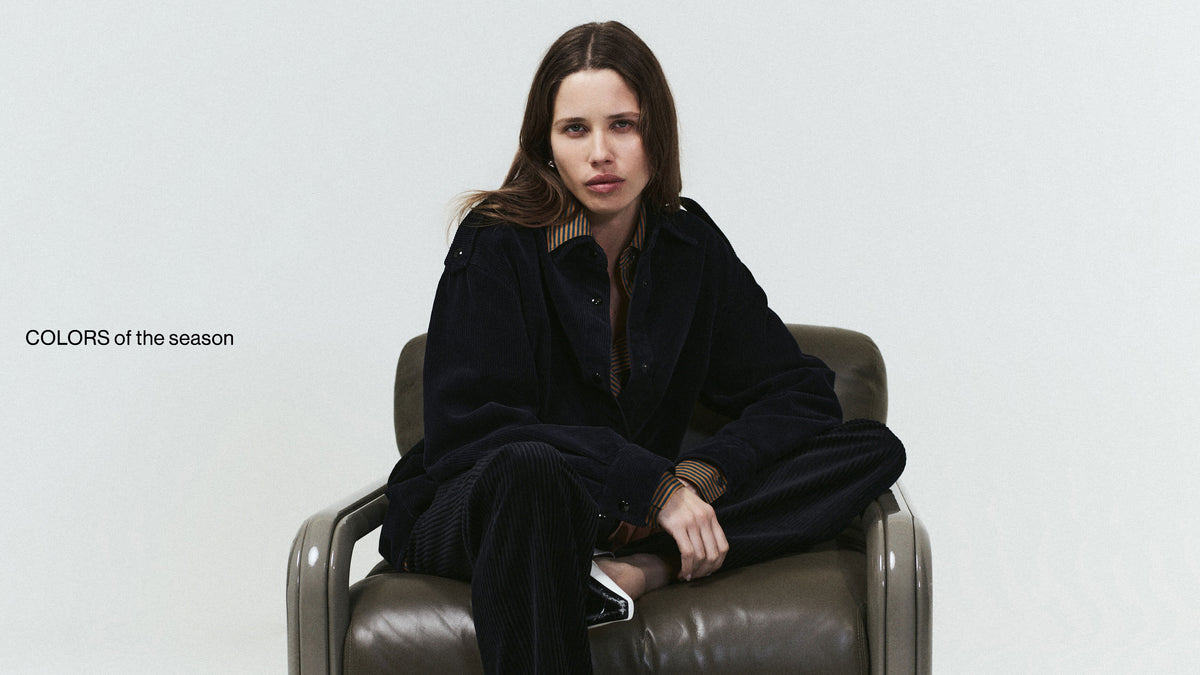 A woman sits in a brown leather armchair wearing a dark navy coat and matching dark trousers against a light gray studio background. Small text in the lower left reads 'COLORS of the season'.