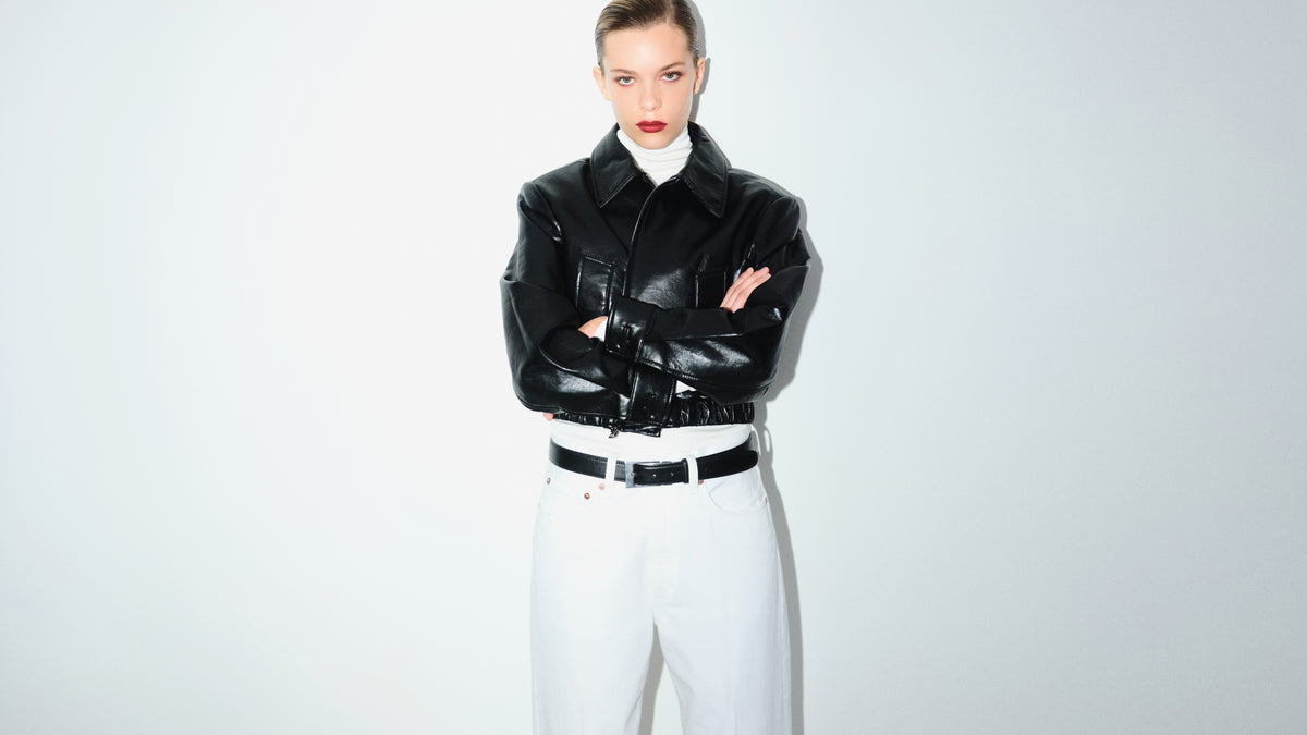 A model stands with arms crossed wearing a glossy black leather jacket over a white turtleneck, paired with white trousers and a black belt, against a plain light gray studio background.