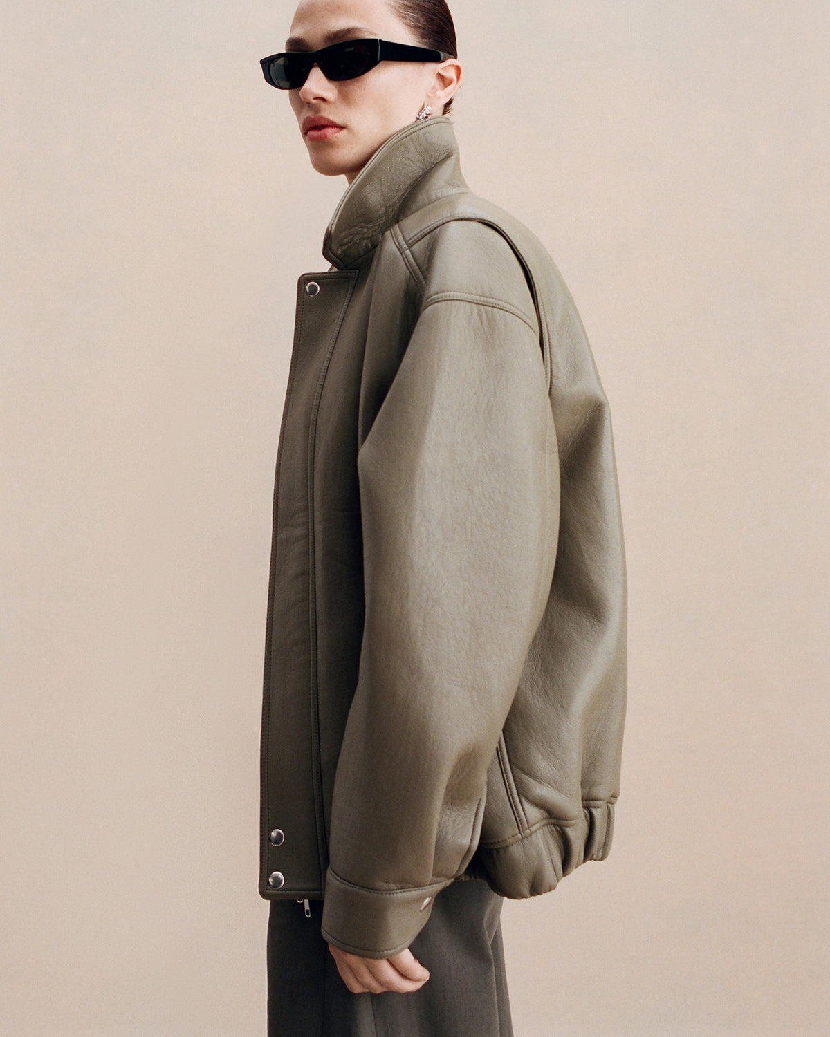A model wearing an oversized olive green leather jacket with a high collar and silver snap buttons, styled with black sunglasses against a solid beige background.