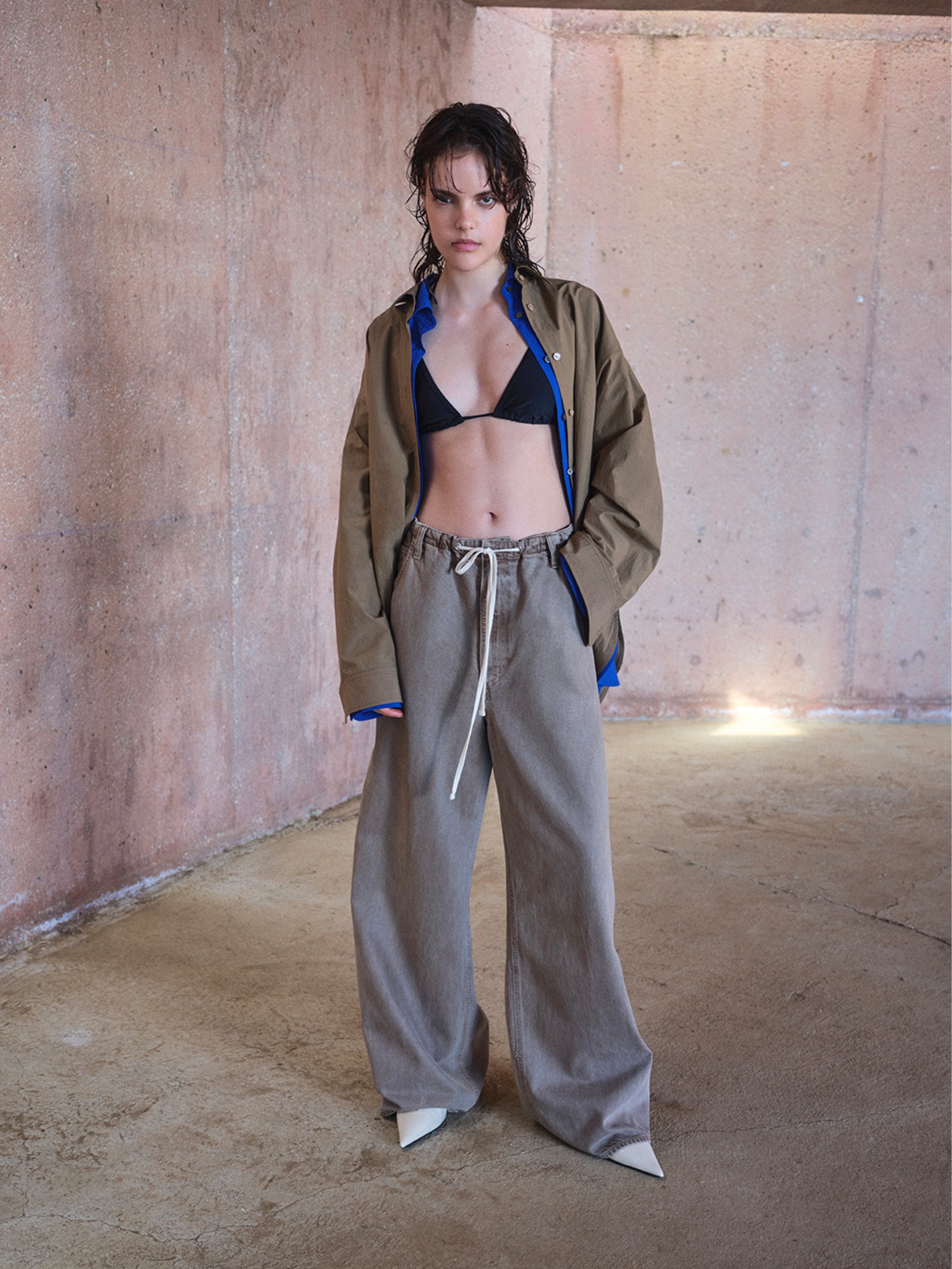 Person wearing a beige jacket, black bra, and gray pants in an industrial setting