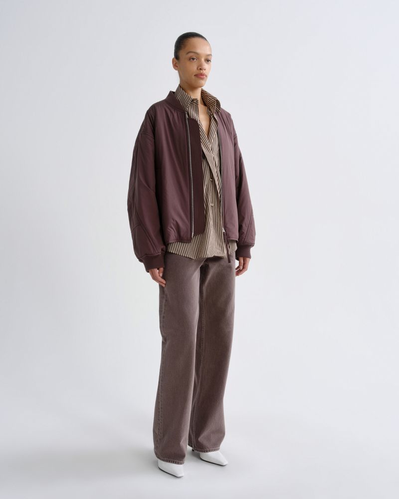 A model stands against a light gray studio background wearing a dark brownish-purple bomber jacket over a beige striped shirt, paired with matching wide-leg trousers and white pointed shoes.