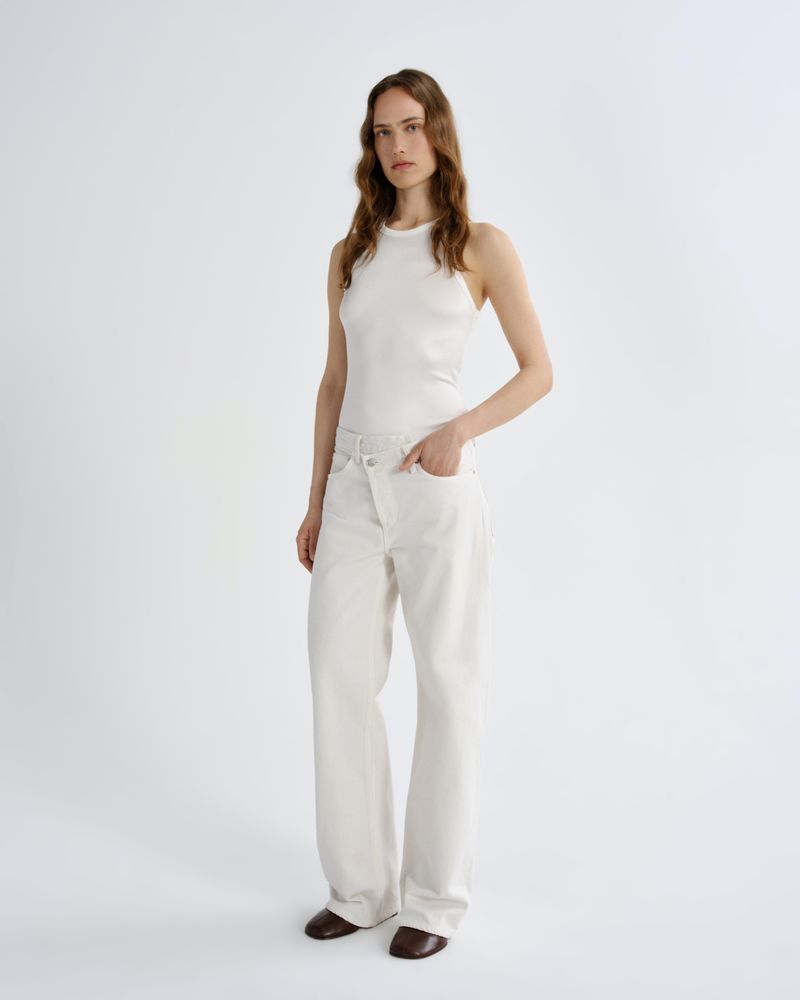 A woman modeling a white sleeveless mock-neck top and matching white wide-leg trousers, standing against a plain light gray studio background.