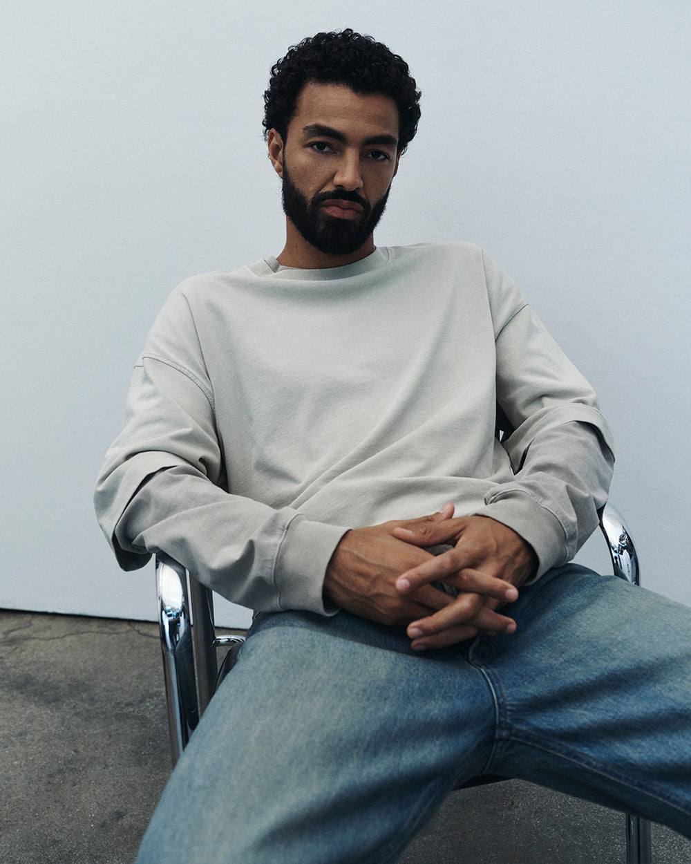 A man with a beard and curly hair sits in a metal chair wearing a light gray crewneck sweatshirt and blue jeans against a plain light gray wall.