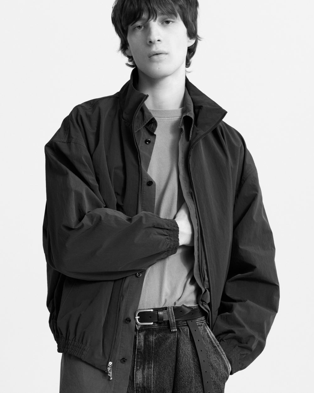 A model wearing a black lightweight jacket over a light gray button-up shirt and dark jeans, photographed in black and white against a plain light background.