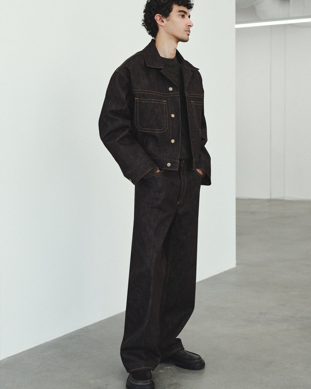 A man wearing a dark brown wide-leg denim jacket and matching wide-leg denim pants, standing in a minimalist white studio space.