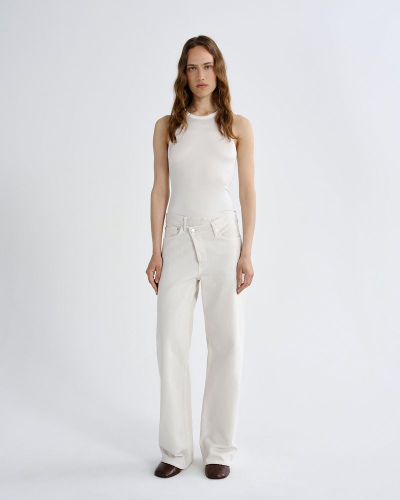 A model stands against a light gray studio background wearing a sleeveless white top and matching white wide-leg pants, with dark brown pointed shoes.