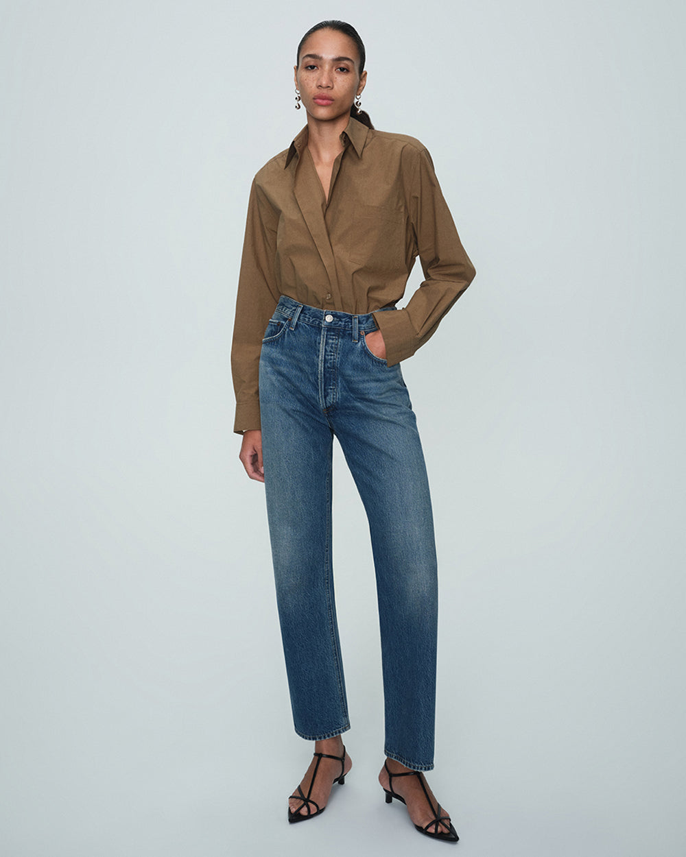 A model stands against a light gray studio background wearing a brown button-up shirt tucked into high-waisted blue jeans, paired with black strappy heels.