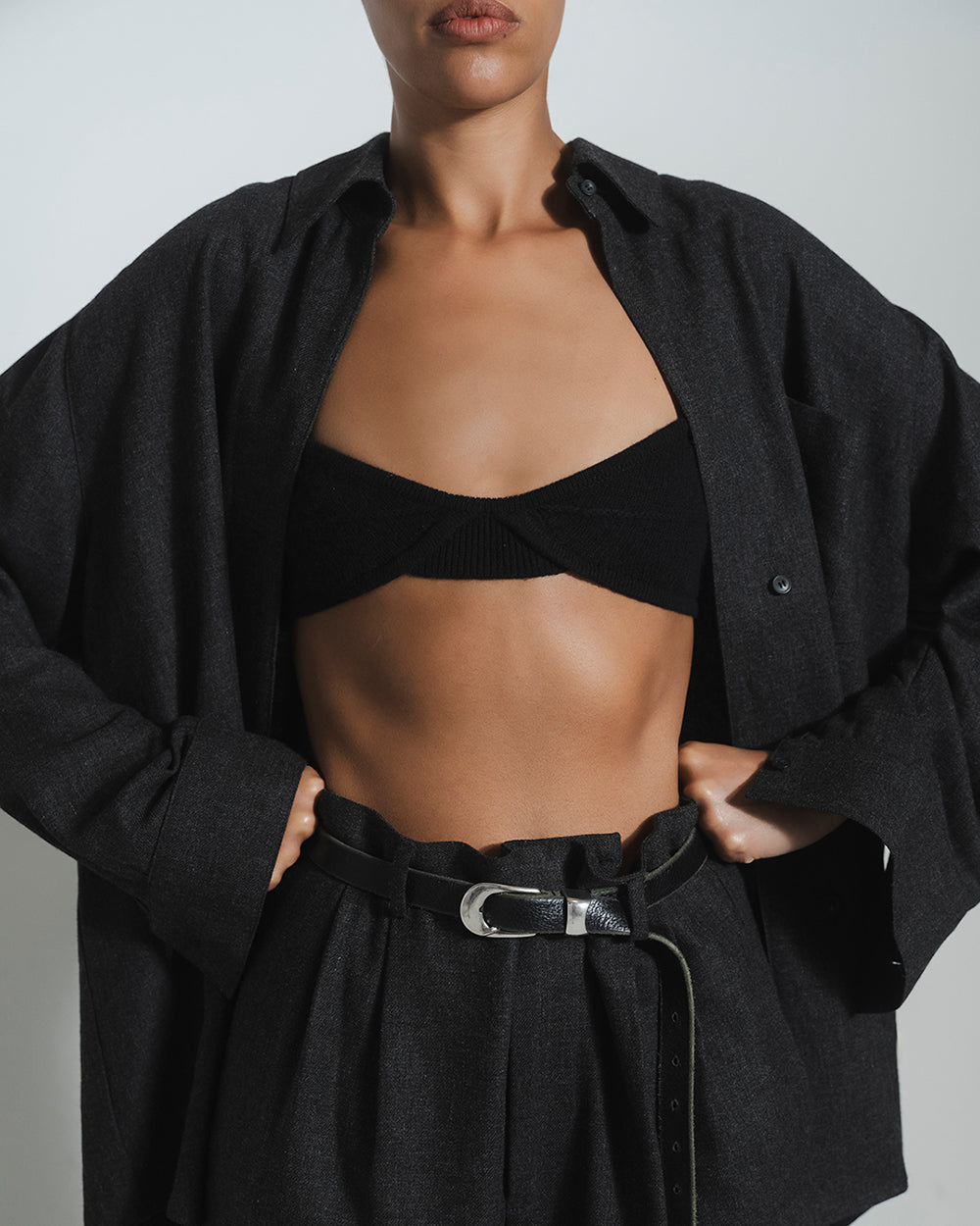A close-up of a model wearing a black ribbed bandeau crop top under an oversized dark charcoal button-up shirt, paired with a black belt and pleated skirt.