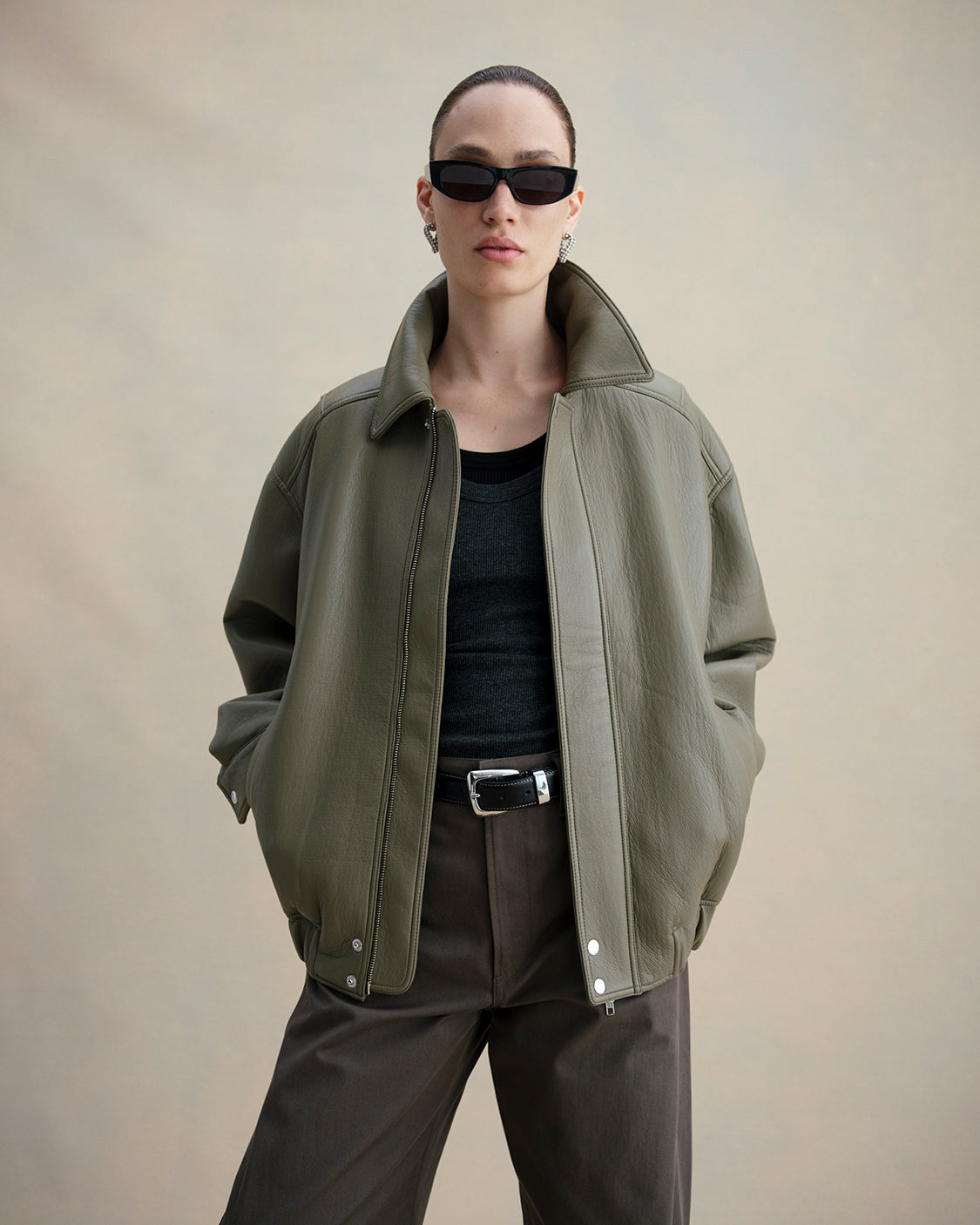 A model wearing an oversized olive green leather jacket, black sunglasses, a black top, and dark brown trousers, standing against a neutral beige background.