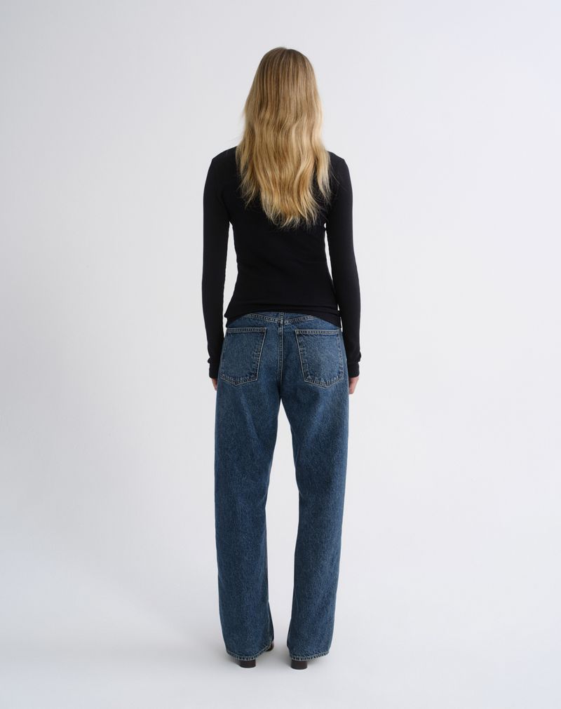 A woman viewed from behind wearing a black long-sleeve top and wide-leg blue jeans against a plain light gray studio background.