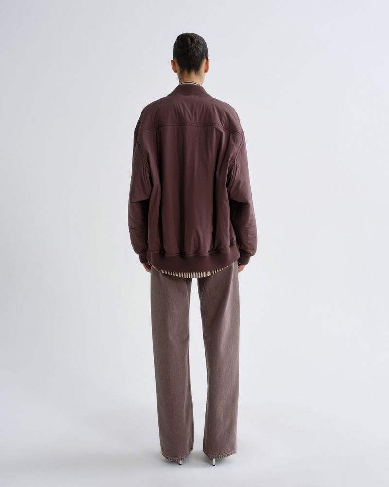 A model viewed from the back wearing a dark brownish-purple bomber jacket and matching wide-leg trousers against a light gray studio background.