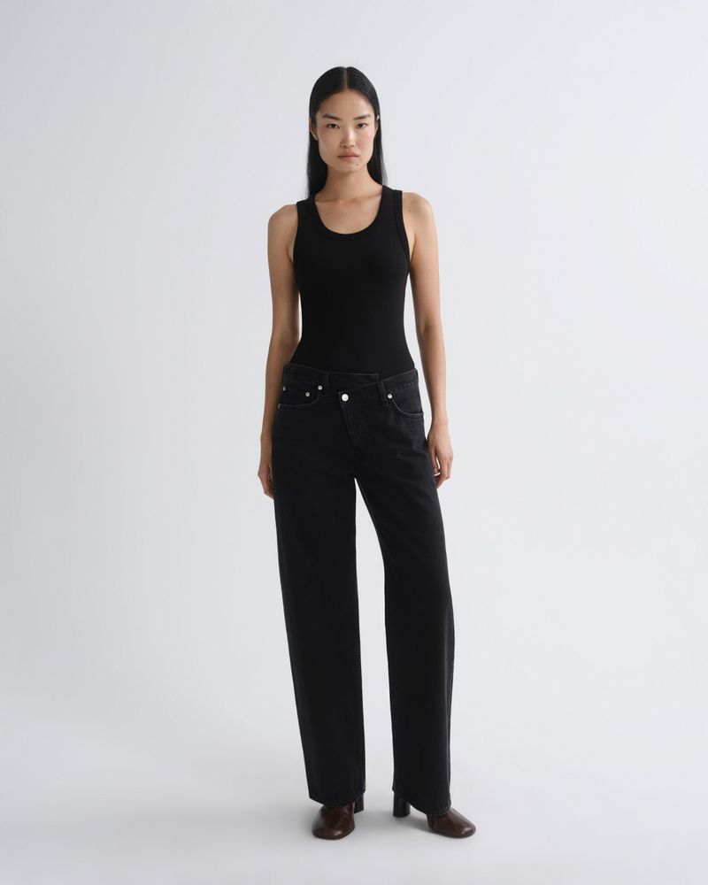 A model stands against a light gray studio background wearing a black sleeveless top and black wide-leg jeans, paired with dark brown shoes.